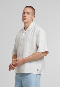 Lahka lanena srajca s kratkimi rokavi v off-white barvi, ki ima izrezan ovratnik, zapenjanje na gumbe in izšito logo na prsih.