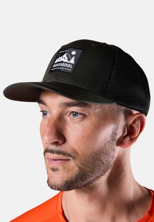 Hombre con gorra de malla Rossignol en verde oscuro y negro con parche de logo de montaña, mirando ligeramente hacia un lado contra un fondo liso.