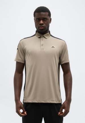 Jonge man die een beige poloshirt met zwarte zijpanelen en een zwarte broek draagt, staand tegen een effen lichte achtergrond.