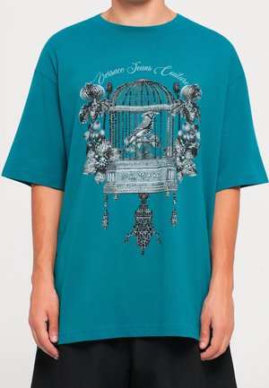 Maglietta color teal con un design dettagliato in bianco e nero di una gabbia per uccelli, un uccello e fiori, e il testo "Versace Jeans Couture" sopra.