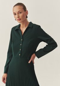 Vestido verde oscuro de manga larga con cuello tipo camisa, detalle de botones y diseño de falda plisada, con tejido suave y acentos dorados.