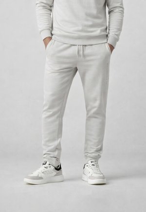 Uomo che indossa pantaloni della tuta grigio chiaro e scarpe da ginnastica bianche, con le mani nelle tasche, in piedi davanti a uno sfondo grigio chiaro uniforme.