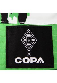 Zwarte stoffen label met wit logo met "B" en "COPA," bevestigd aan een groen-witte geprinte achtergrond.