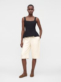 Femme debout portant un haut noir sans manches, un short crème jusqu'aux genoux et des chaussures plates marron à lacets sur un fond clair uni.