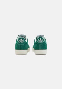 Os ténis de camurça verde apresentam uma sola de borracha branca, um tab de calcanhar em contraste e o logótipo "Stan Smith" com o trevo da adidas na parte de trás.