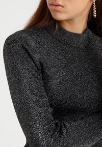 Pull noir scintillant avec un col haut et des manches longues. Le tissu est texturé avec des fils métalliques, créant un effet étincelant.
