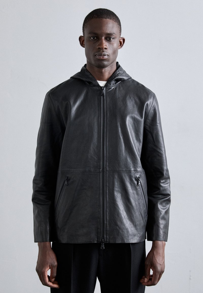 Casaco de couro preto com capuz, apresentando um fecho frontal, dois bolsos laterais e uma textura suave. Design minimalista com linhas limpas.