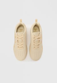 Beige joggesko med glatt syntetisk materiale, snøring, perforeringer for ventilasjon og diskrete trykte detaljer.