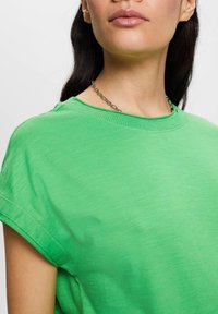T-shirt in cotone verde con maniche corte, collo rotondo e una texture liscia. Caratterizzata da cuciture visibili lungo il collo e l'orlo.