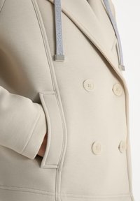 Gros plan sur un manteau beige à double boutonnage avec de gros boutons, une poche latérale et des cordons gris avec des embouts métalliques portant l'inscription « DISCOVER NEVER ».