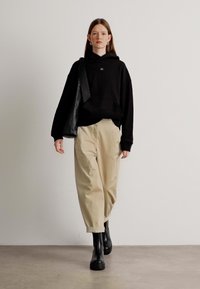 Felpa nera oversize con logo, pantaloni beige larghi e stivaletti neri alla caviglia. Il modello tiene una borsa tote nera, in piedi su uno sfondo neutro.