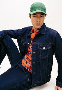 Τζιν μπουφάν σε σκούρο μπλε με πορτοκαλί και ριγέ πουκάμισο από κάτω. Το πράσινο καπέλο έχει λογότυπο. Ορατές λεπτομέρειες κουμπιών και τσέπης.