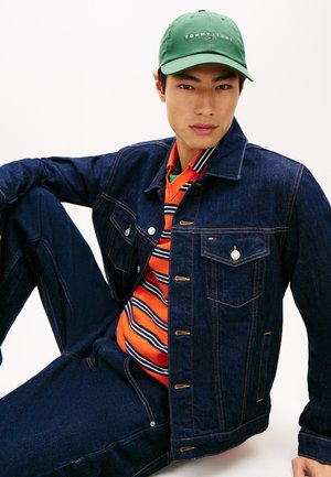 Kurtka dżinsowa w ciemnym niebieskim kolorze z pomarańczową i paskowaną koszulą pod spodem. Zielona czapka z logo. Widoczne detale guzików i kieszeni.