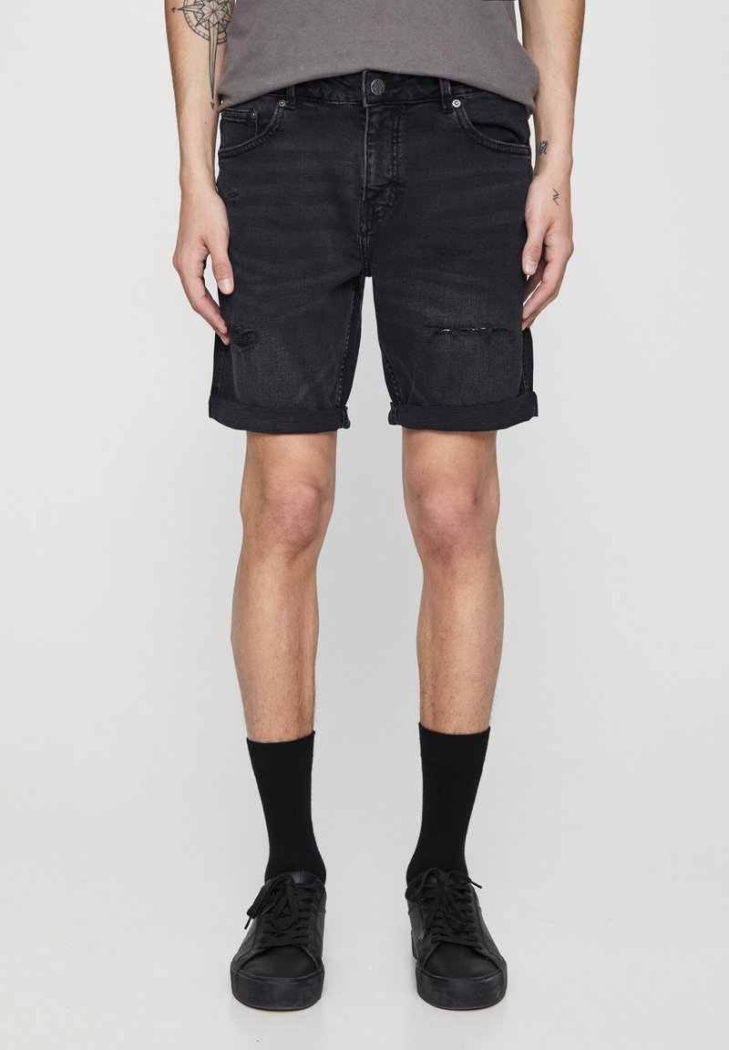Shorts en denim noir avec ourlets roulés, présentant des détails usés et une coupe ajustée. Assortis avec des chaussettes noires et des baskets.