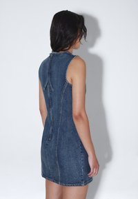 Denimmekko, jossa on korkea kaulus, hihaton muotoilu ja takana piilotettu vetoketju sekä nauha-yksityiskohta. Rikkoutunut ompelu korostaa.