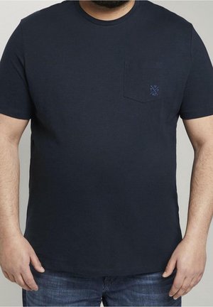 Mand iført en mørk navy kortærmet T-shirt med en lille lomme og et emblem på venstre bryst, parret med blå jeans.