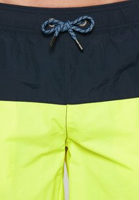 Uimashortsit, joiden yläosa on tummansininen ja alaosa kirkkaan neonkeltaisena. Sisältää kiristysnauhan, jossa on sinisiä ja mustia kuvioituja naruja.