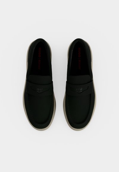 Paire de mocassins noirs sans lacets avec semelles beiges, logo "HUGO" embossé sur le dessus, vus de dessus sur fond blanc.