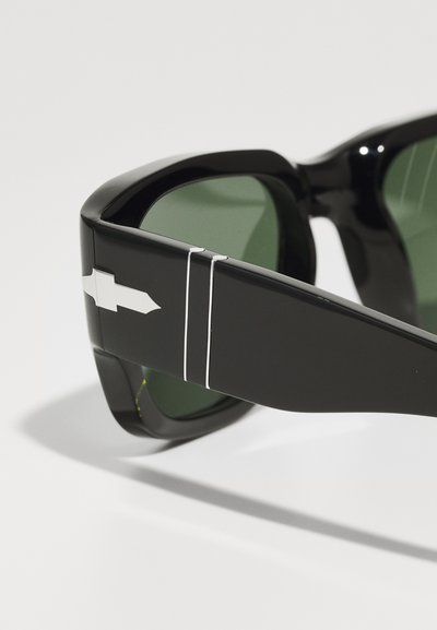 Črne sončne očala z zelenimi lečami, ki imajo sijajno površino, široke okvirje in srebrno puščico na strani.