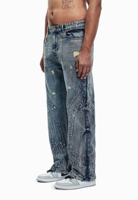 Φαρδιά μπλε τζιν παντελόνια με φθαρμένες λεπτομέρειες, γεωμετρικό σχέδιο στα πλαϊνά και εμφανείς ραφές. Φοριούνται με αθλητικά παπούτσια.