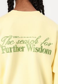 Yellow sweatshirt featuring green embroidered text: "The search for Further Wisdom" with brand details above in a smaller font.