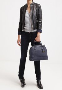 Osoba w czarnej skórzanej kurtce, szarym swetrze, ciemnych dżinsach i czarnych butach, trzymająca w dłoniach torebkę w fakturze w kolorze granatowym.