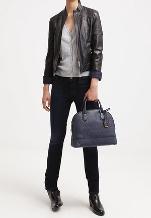 Persona con giacca di pelle nera, cardigan grigio, jeans scuri, stivali neri, che tiene una borsa a tracolla blu navy con texture per le maniglie.