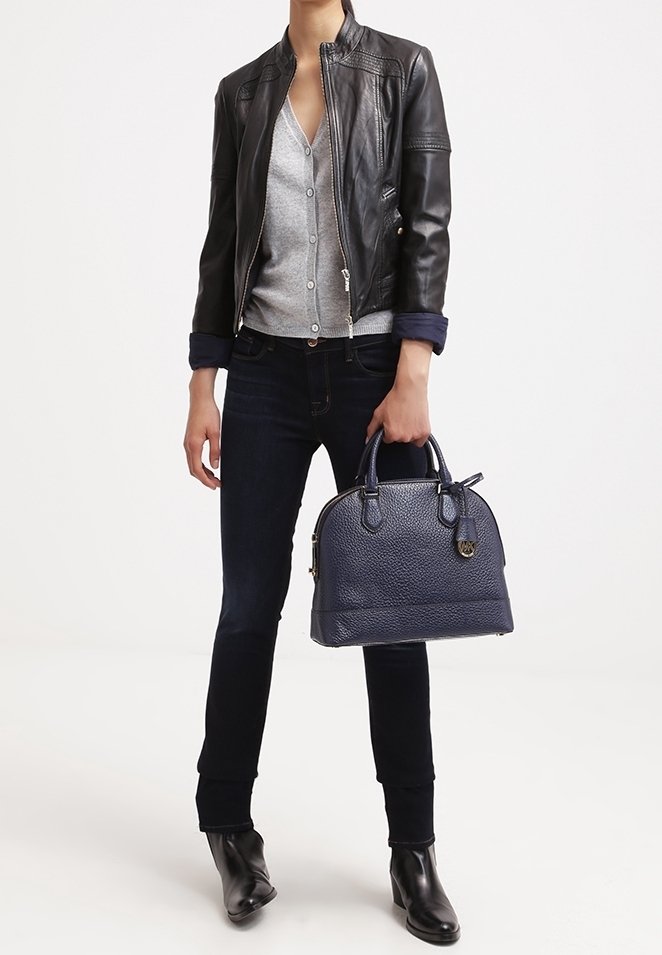 Osoba w czarnej skórzanej kurtce, szarym swetrze, ciemnych dżinsach i czarnych butach, trzymająca w dłoniach torebkę w fakturze w kolorze granatowym.