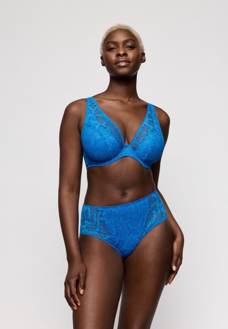 Femme aux cheveux blonds courts portant un ensemble assorti soutien-gorge en dentelle bleu vif et culotte taille haute, debout devant un fond uni.