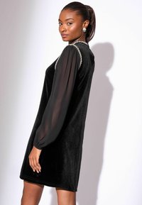 Mustast sametist kleit läbipaistvate läikivate varrukatega, kaunistatud kaelusega, põlveni ulatuva lõikega, siledast tekstuurist ja lihtsa siluetiga.