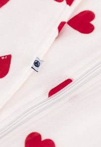 Βαμβακερά κλινοσκεπάσματα με λευκό φόντο που διαθέτουν κόκκινα σχέδια καρδιών και λευκό φερμουάρ. Περιλαμβάνει μια μικρή ετικέτα μάρκας στη γωνία.