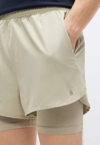 Pantaloni scurți atletici de culoare bej deschis, cu talie elastică, buzunar lateral și strat interior fixat. Textură moale și detaliu minimal cu logo pe tiv.