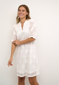 Biele čipkované šaty s kvetinovým vzorom, krátkymi puff rukávmi, výstrihom do V a uvoľneným strihom, ktorý zobrazuje zložitú detailnosť a štruktúru.