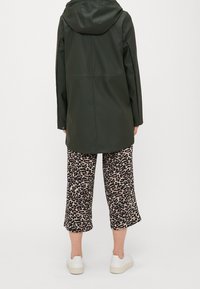 Ciemnozielona wodoodporna parka z kapturem, długimi rękawami i prostą sylwetką, zestawiona z szerokimi spodniami w panterkę i białymi butami.