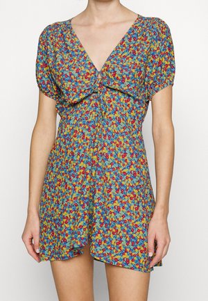 Femme portant une robe courte multicolore à fleurs avec des manches bouffantes et un détail de nœud à l'avant au niveau de la poitrine.