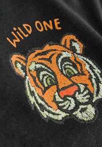 Čierna tkanina s vyšitým dizajnom tigra s oranžovou a bielou farbou, zelené oči, doplnená textom "WILD ONE" nad tým.