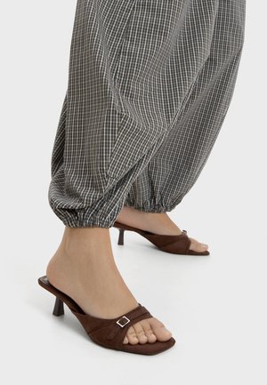 Picioare purtând pantofi maro cu toc mic și deschiși în față, cu o cataramă mică, asortați cu pantaloni largi maro și albi în carouri, cu manșete elastice.