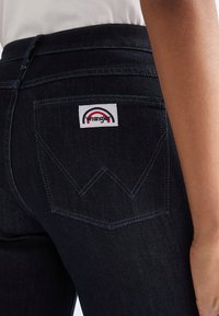 Close-up of dark blue denim jeans back pocket with white Wrangler label featuring red and black semi-circle logo on person wearing white top.