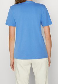 Modré bavlněné tričko s krátkým rukávem, kulatým výstřihem a hladkou texturou. Zpětný pohled neukazuje žádné vzory ani viditelné loga. V kombinaci se světle barevnými kalhotami.