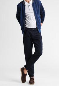 Mężczyzna w granatowej kurtce, białej koszuli polo, ciemnych spodniach i brązowych butach, stoi z nogami skrzyżowanymi i rękami w kieszeniach.