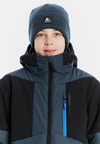 Blaue und schwarze isolierte Jacke mit hohem Kragen, Reißverschlusstasche und Logo-Detail. Getragen mit einer passenden blauen Strickmütze.