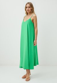 Eine Frau steht in einem einfachen, leuchtend grünen Kleid mit dünnen Trägern und trägt beigefarbene Sandalen vor einem schlichten weißen Hintergrund.