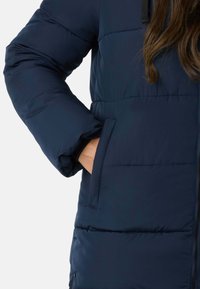 Marineblå quilted pufjakke med lynlås foran og sidelommer. Stoffet ser glat og isoleret ud.