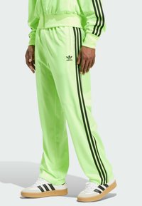 Neon green tracksuit pants with black side stripes and an embroidered logo. Made of smooth, shiny material with a relaxed fit.