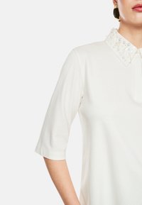 Weißes Poloshirt mit strukturiertem Kragen, verziert mit dekorativen Perlenakzenten und kurzen Ärmeln. Weicher, glatter Stoff mit lockerer Passform.