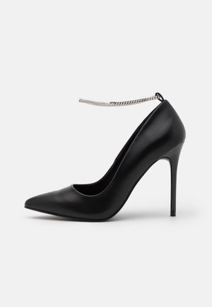 Schwarzer High-Heel-Pumps aus glattem Leder, mit spitz zulaufender Zehenpartie und dünnem Knöchelriemen mit metallischer Kettenverzierung.