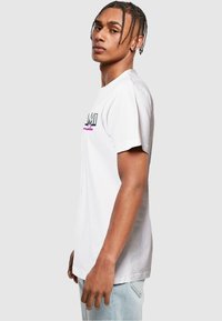 Tricou alb din bumbac cu mâneci scurte, având un grafic multicolor "MIAMI" pe piept, purtat cu blugi de culoare albastră deschisă.