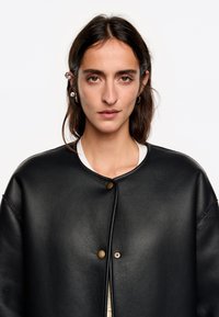 Chaqueta de cuero negra con cuello redondo, que presenta un cierre de botón único y mangas anchas. Textura suave con un diseño minimalista.