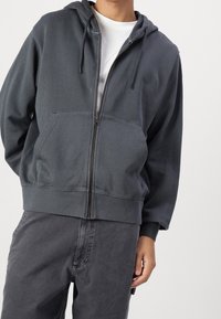 Felpa con zip di colore grigio scuro, dotata di tasca frontale, cappuccio con coulisse e orlo a coste. Realizzata in un tessuto morbido con una superficie liscia.