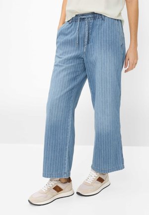 Person iført blå denim bukser med brede ben og elastisk talje og beige sneakers med brune detaljer.